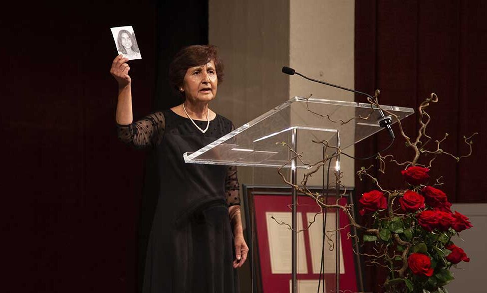 A woman on stage at a lectern speaks while holding up a black-and-white photo of Mona Mahmoudnejad, age 17.