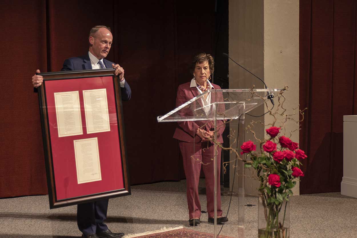 A man stands behind a lectern and holds up a large framed wall hanging that contains three pages of a document, as a woman stands nearby, both on a stage with an arrangement of roses at the front.