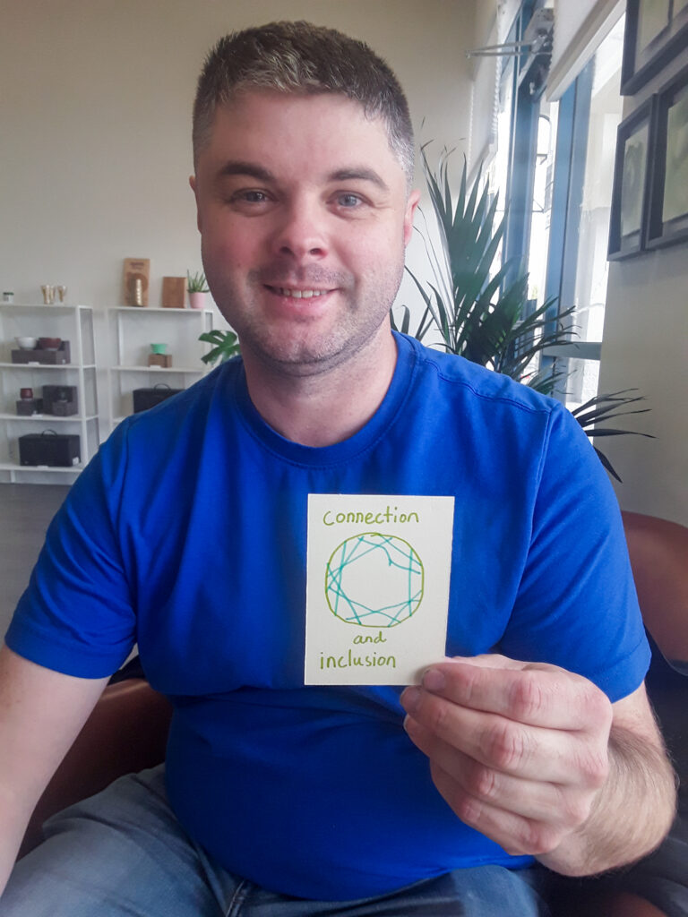 A smiling white skinned man sits holding his artwork that says "Connection and Inclusion"