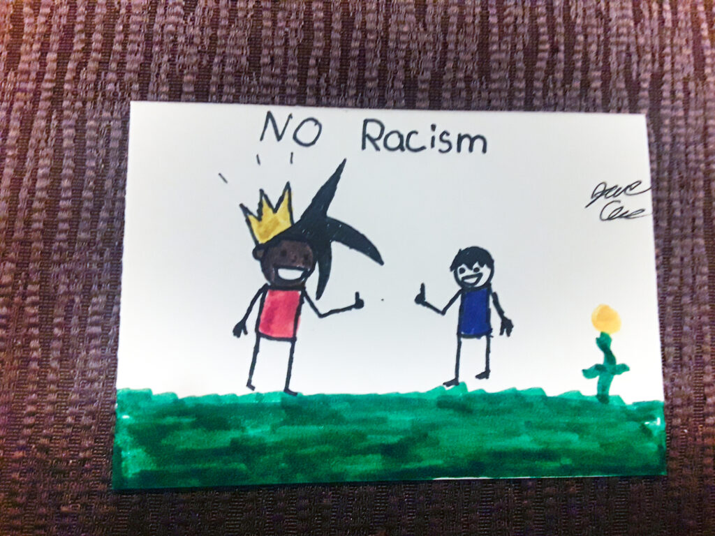 A piece of small drawing that shows a black child and a white child with their thumbs up under the words "No Racism"
