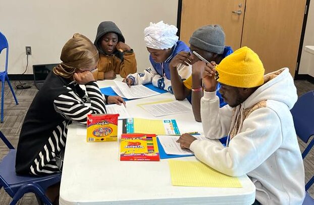 A group of young African people sit at a table studying and creating art.