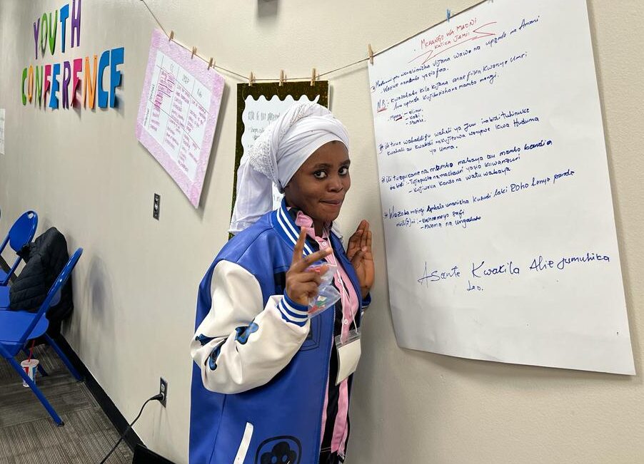 Young African woman stands in front of a white board poster written in Swahili.