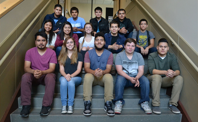 A group of Hispanic and white teenagers sit on school steps and smile at the camera.
