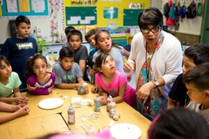 An African American teacher engaged in conversation with a group of middle school-aged Hispanic students, surrounding a table with arts and crafts supplies.